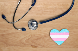 A heart painted with the colors of the trans flag and a stethoscope sit on a table.