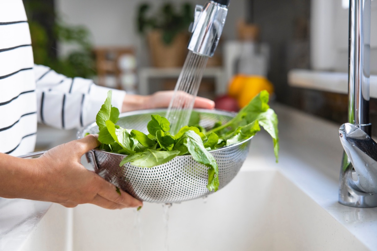 A person washing arugula salad greens by holding a silver metal colander in their hands over a white sink as water flows from a faucet, passes over the greens, and then drips from the bottom of the colander.