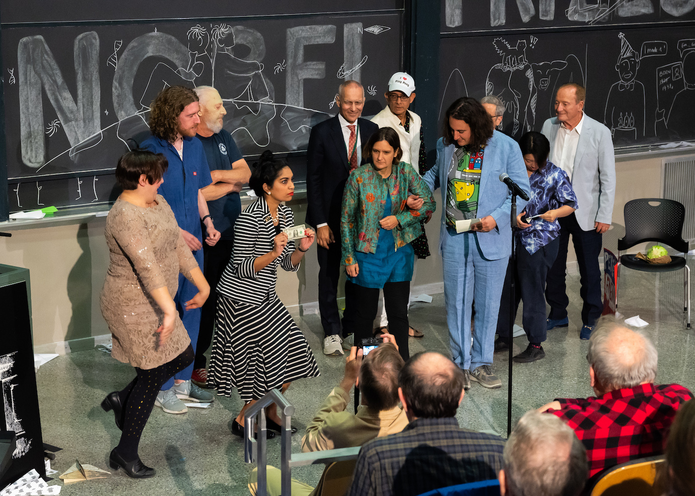 A woman holds a dollar bill on a stage with several others in expressive outfits stand by her.