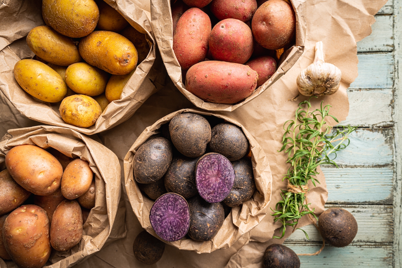 Various varieties of new colorful, white, red and purple potatoes in paper bags on white wooden background, top view