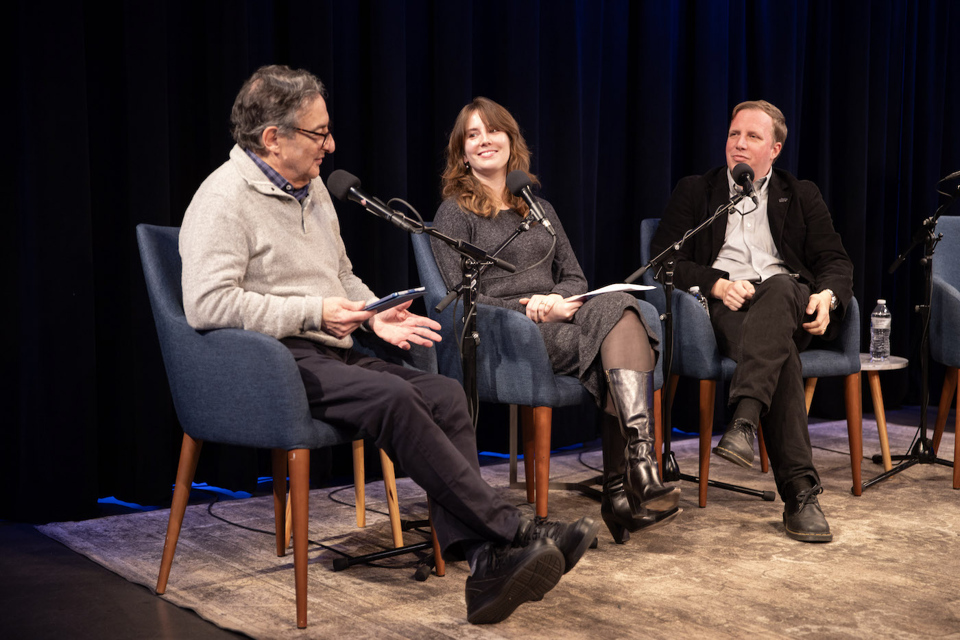 Three people have a conversation into microphones on stage.