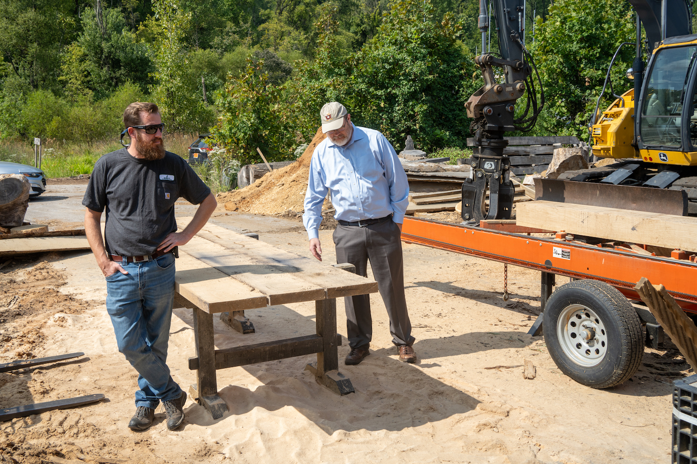 Two men look at a wooden table in a sandy outdoor work area.