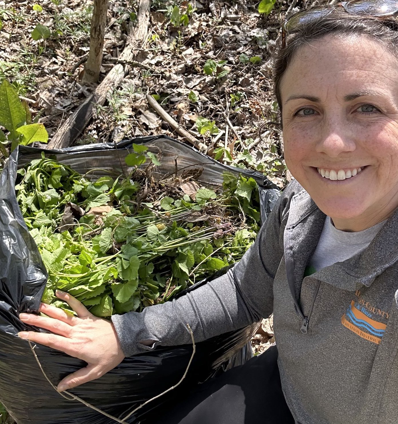 A woman smiling next to a trash bag filled with green weeds.