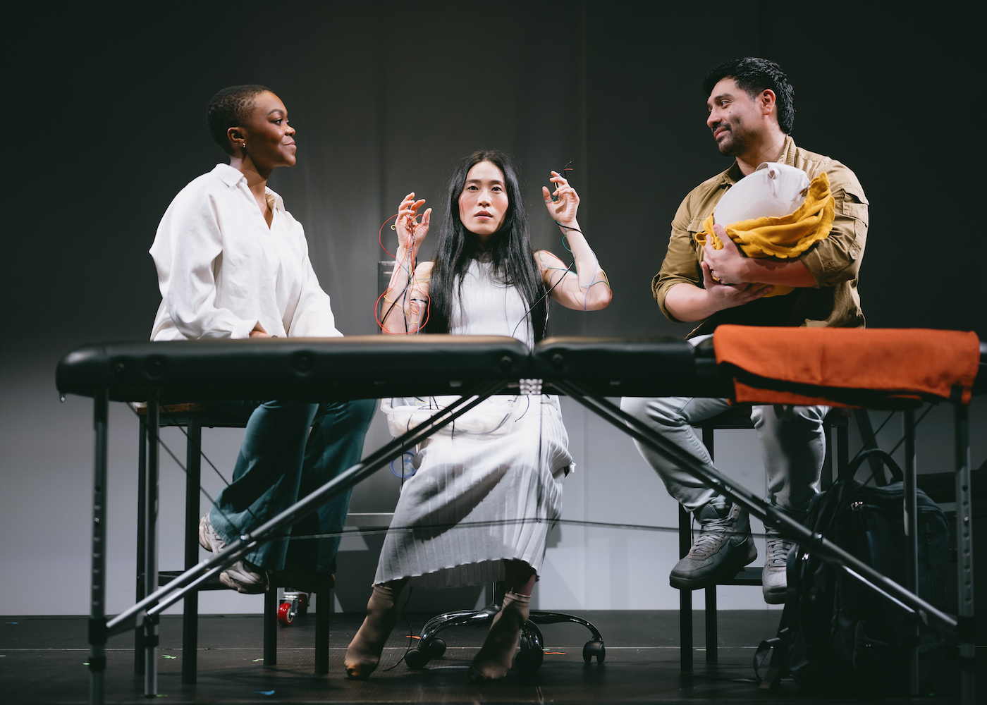 Three people sit on a stage, performing, The woman in the center is hooked up to a bunch of wires, and the man to the right is holding something like a baby