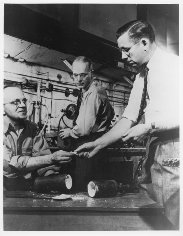 A few people passing items between their hands. On the table in front of them is a capsule filled with spilling white powder