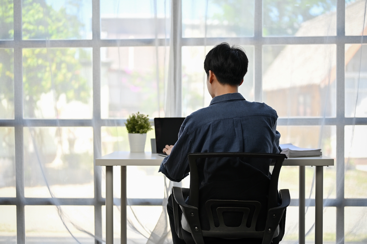 Back view image of a focused businessman working on his work on his laptop computer at his desk by the window in a modern office.