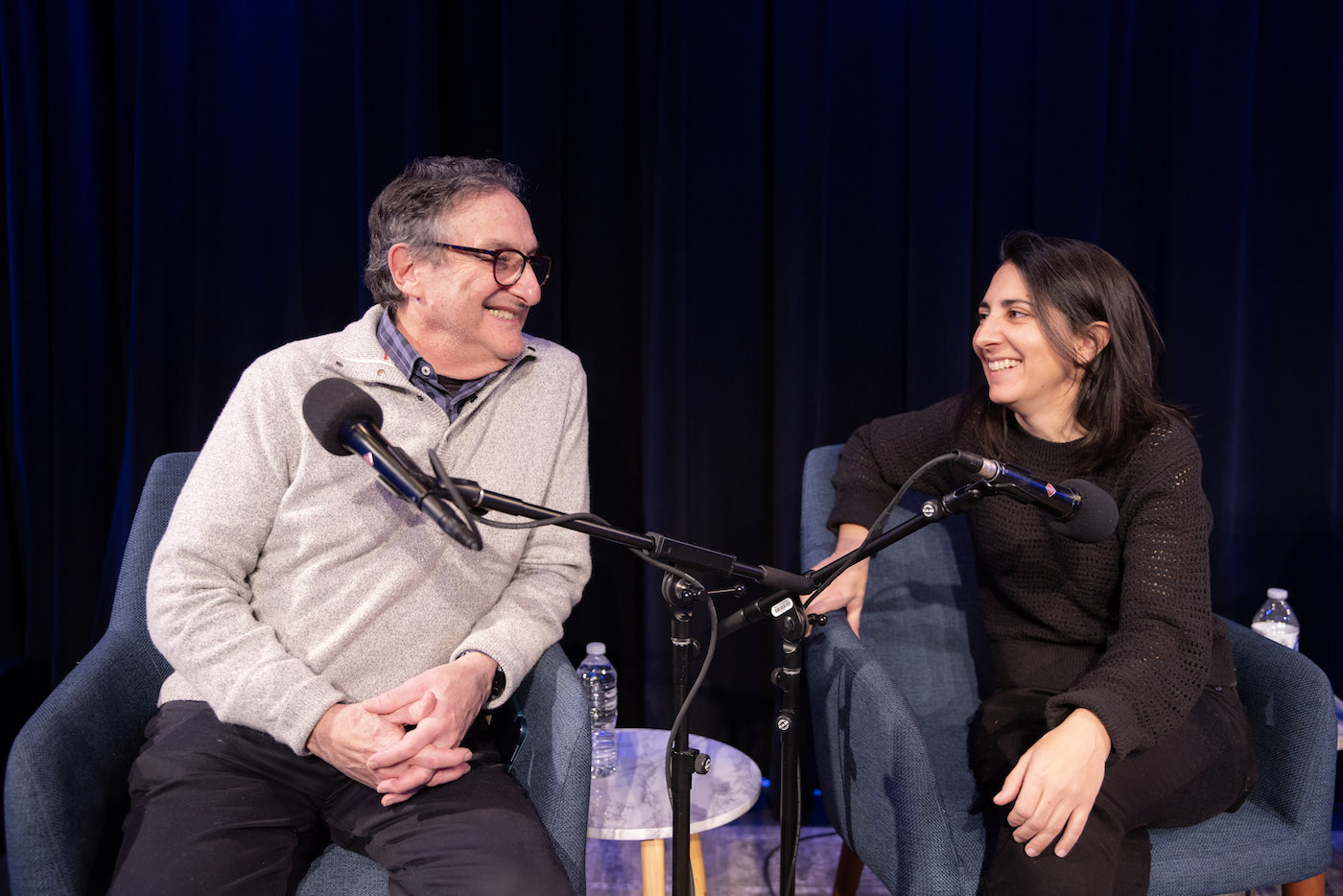 Ira Flatow and Flora Lichtman sitting in front of microphones, smiling at each other.