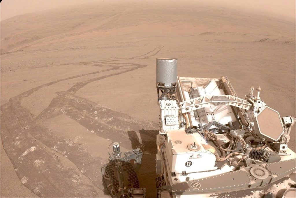 A mars rover on the dusty, pinkish desert surface of Mars, with its tracks zigzagging into the hazy background.