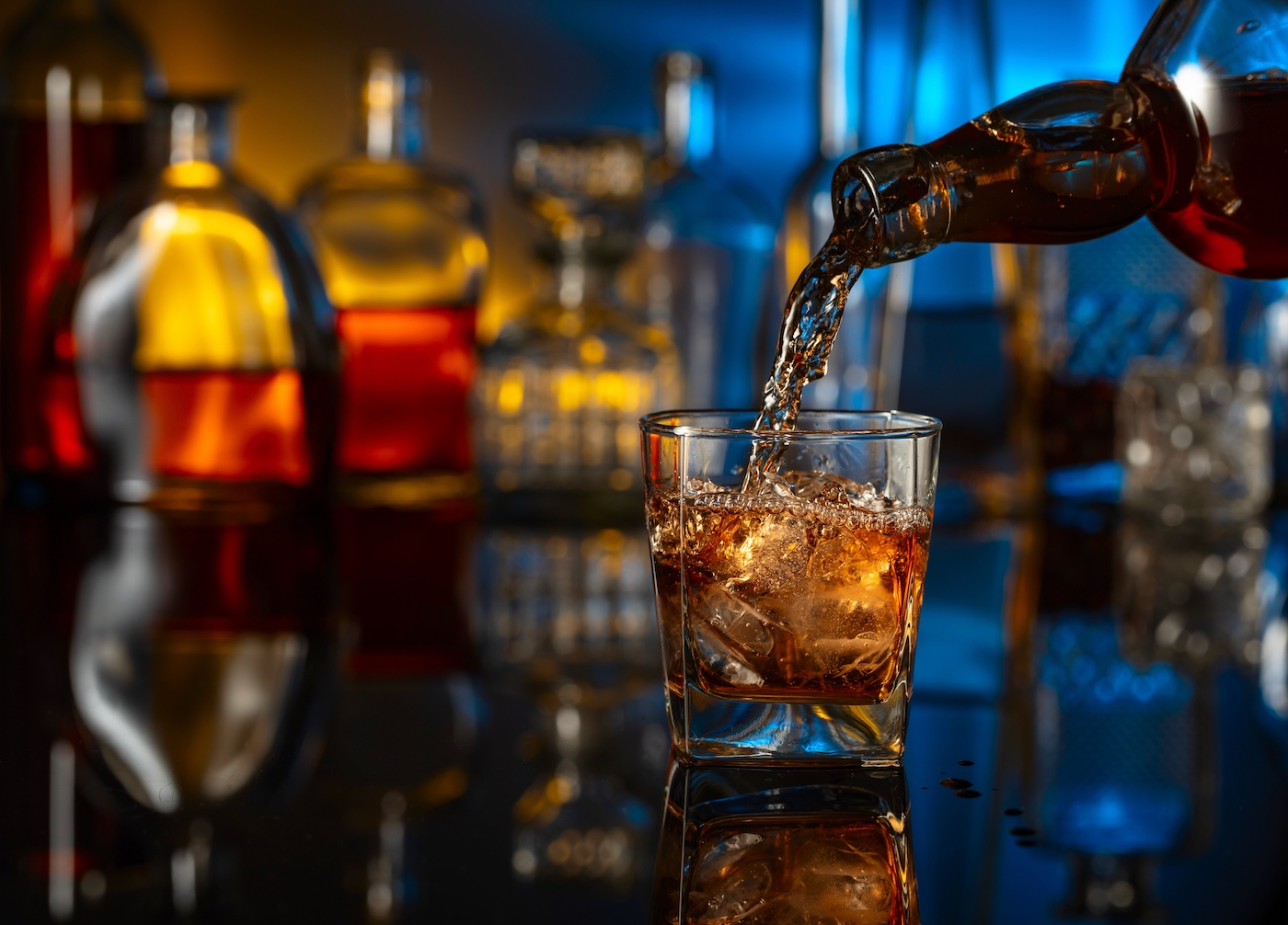 Whiskey is poured from a bottle into a glass with ice. Glass of whiskey on a black reflective background. In the background bottles with various alcoholic drinks.