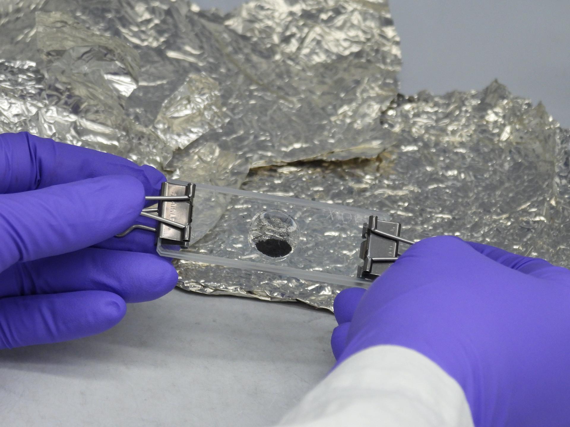 A pair of gloved hands holding a glass slide which contains some black dust in its center