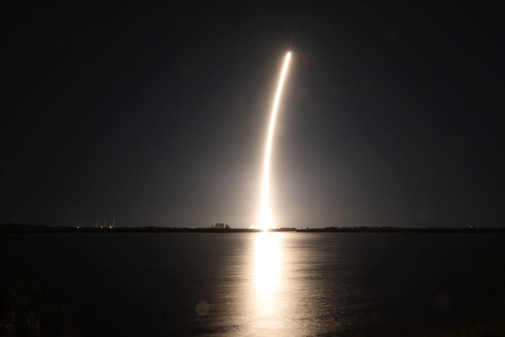 A bright golden streak coming off of a rocket traveling into space, reflected over a large body of water