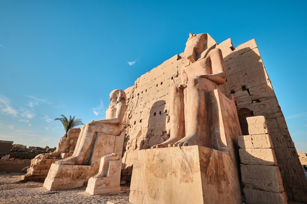 Beige statues of seated pharoahs in front of a boxy structure soar against a blue sky.