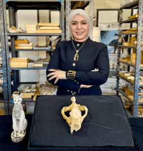 A woman wearing a hijab stands proudly in front of a partial animal skull and a statue of a cat.