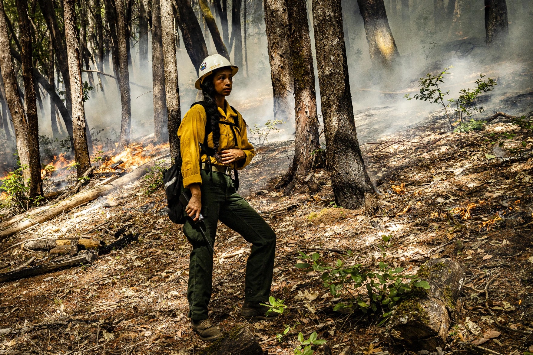 A woman wearing a construction helmet stands holding a radio in a smoky wooded area.