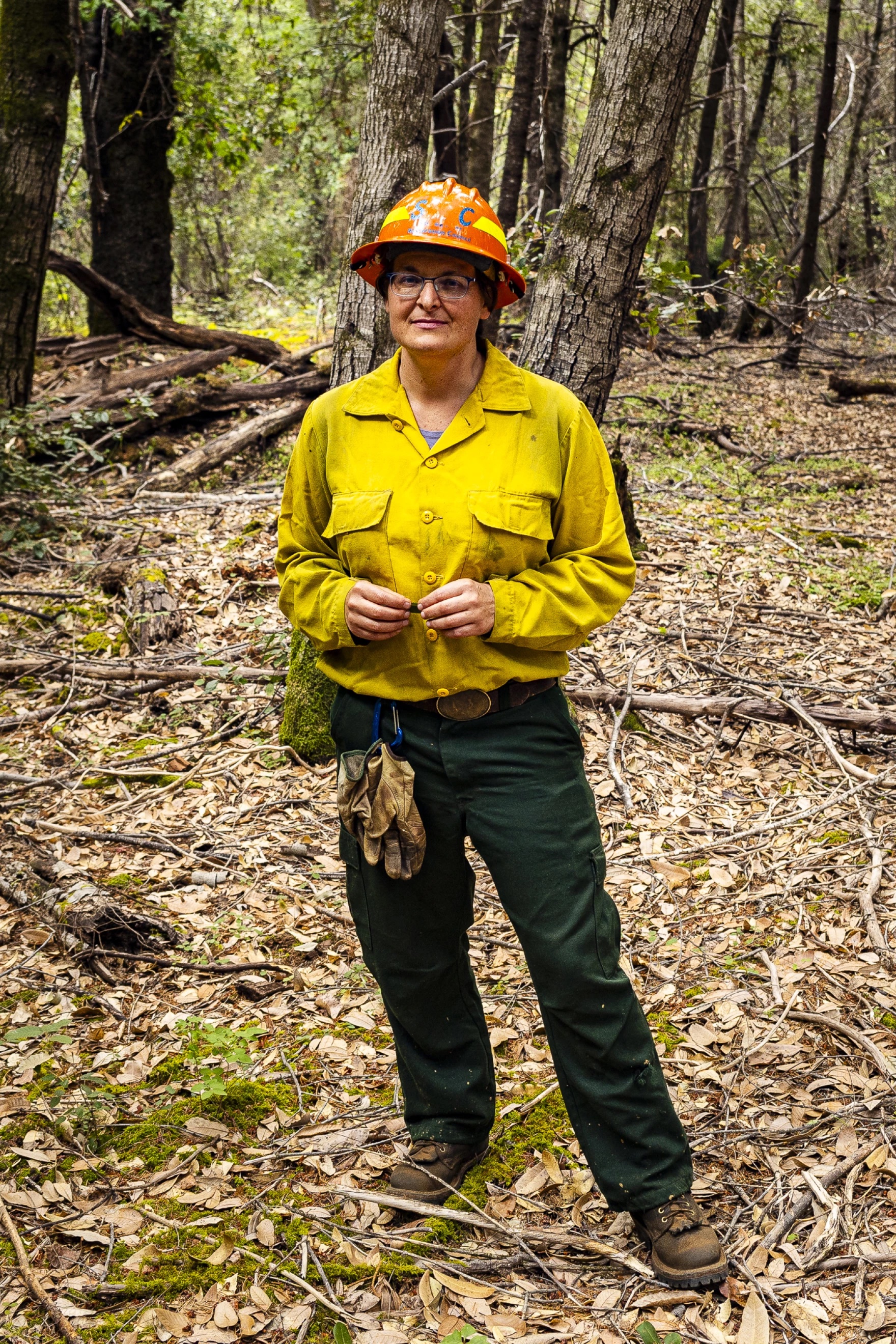 A person wearing a yellow hat and a construction helmet stands in a wooded area