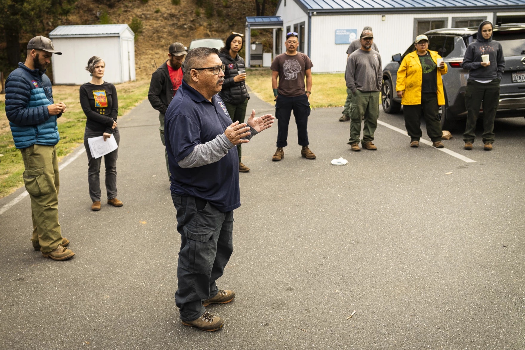 A man speaks to a circle of people outside a small building.