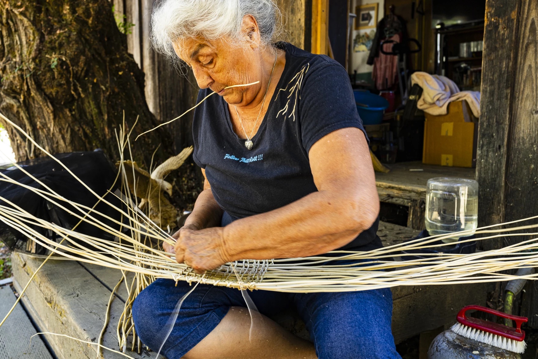 A woman weaves a basket out of long dried plant strands. She holds a stem in her mouth.