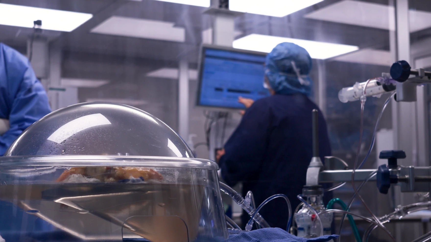 A person in PPE enters information into a computer. In the foreground, an organ rests inside a machine.