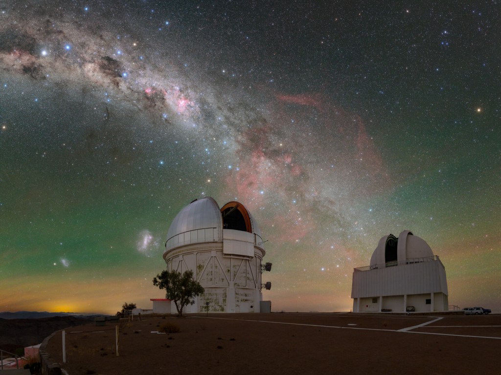 Below the southern skies, the telescopes of Cerro Tololo Inter-American Observatory (CTIO), a Program of NSF NOIRLab, bask in the starlight of the brilliant Milky Way. Speckled with multi-colored stars, our home galaxy in this image stretches down into the city-lit horizon, just grazing past the Víctor M. Blanco 4-meter Telescope (left) and SMARTS 1.5-meter Telescope (right). The Small and Large Magellanic Clouds that accompany our home galaxy are seen to the left of the Blanco telescope. In between the telescopes blooms a deep shade of red that permeates through the tendrils of the Milky Way. This is the Gum Nebula, an emission nebula that blazes with the light of gas ionized by its host stars. From our perspective it holds a bountiful number of cosmic objects, from globular clusters, to the Vela Pulsar, and even to an 11,000-year-old supernova remnant. This photo was taken as part of the recent NOIRLab 2022 Photo Expedition to all the NOIRLab sites.