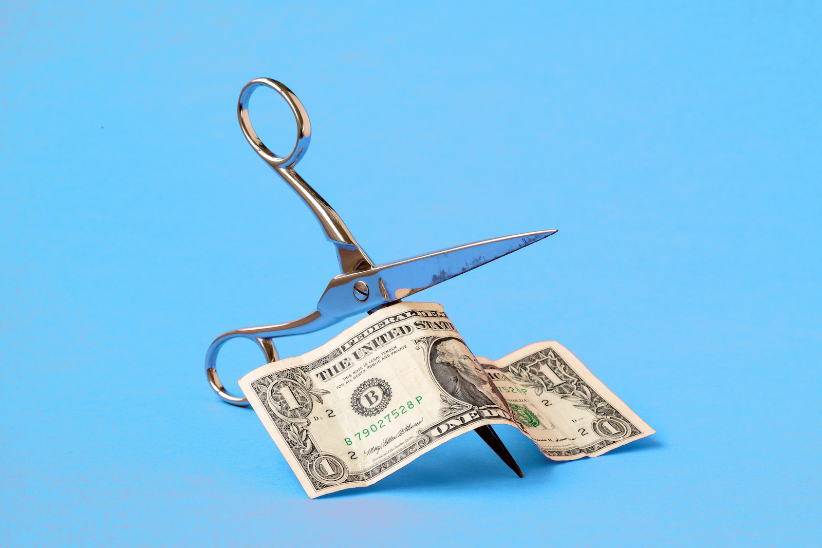 Cutting one dollar with scissors on blue background. Concept on the topic of devaluation of money.