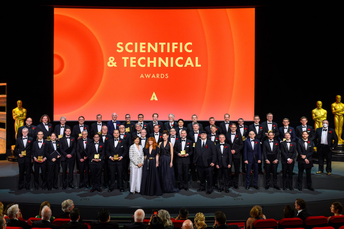 A group of awardees standing in black-tie dress. Actress Natasha Lyonne is at the front, with Academy president Janet Yang. A big sign is in the background that reads "Scientific and Technical Awards"